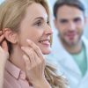 New sensation. Profile of enthusiastic pretty woman with long blond hair touching her hands to ear and man in white coat watching behind in medical office