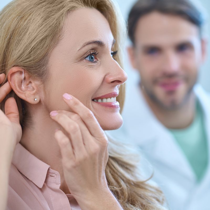 New sensation. Profile of enthusiastic pretty woman with long blond hair touching her hands to ear and man in white coat watching behind in medical office
